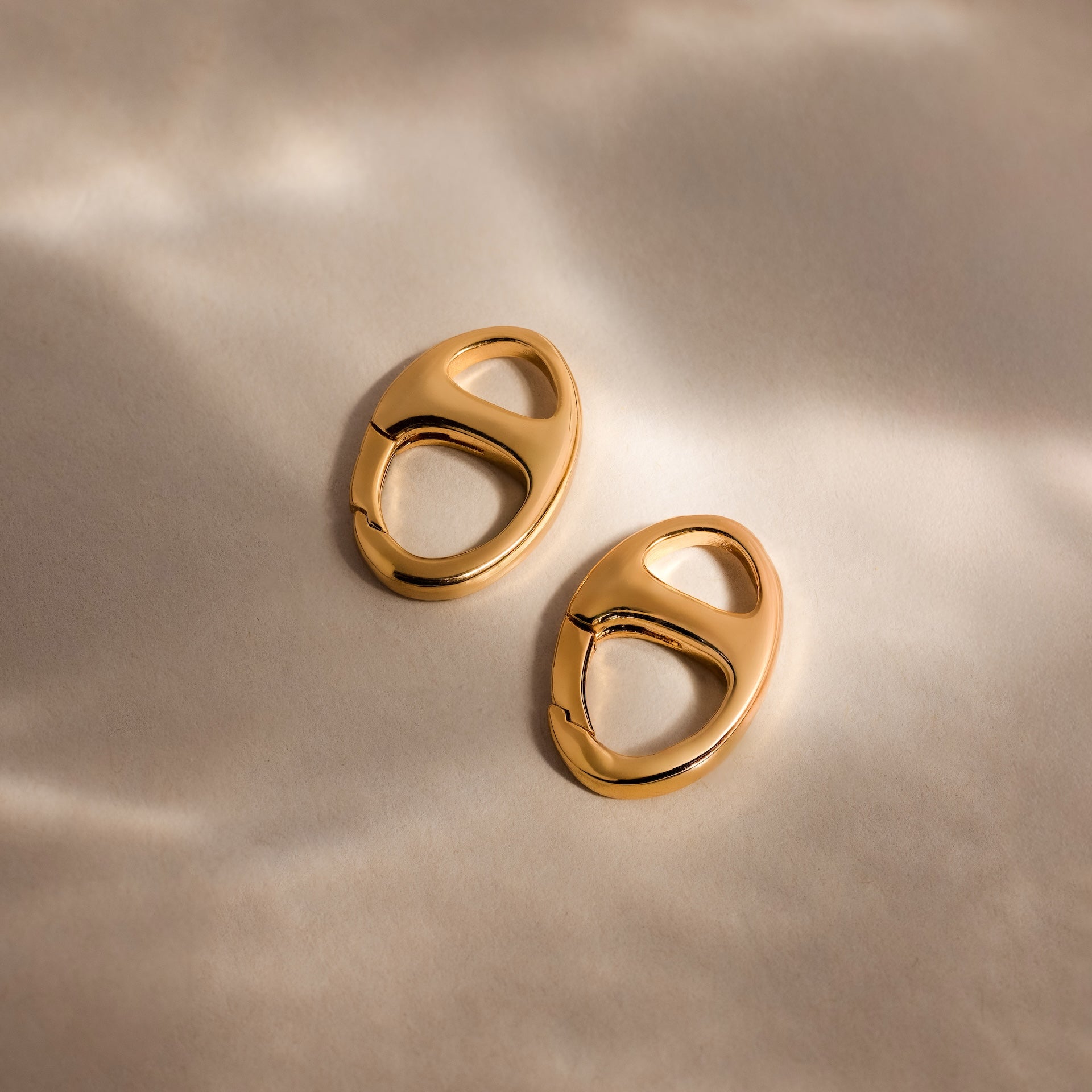 The Connector Clasp: two gold oval hoop earrings with cut-out details rest on a soft, neutral background with gentle shadows.