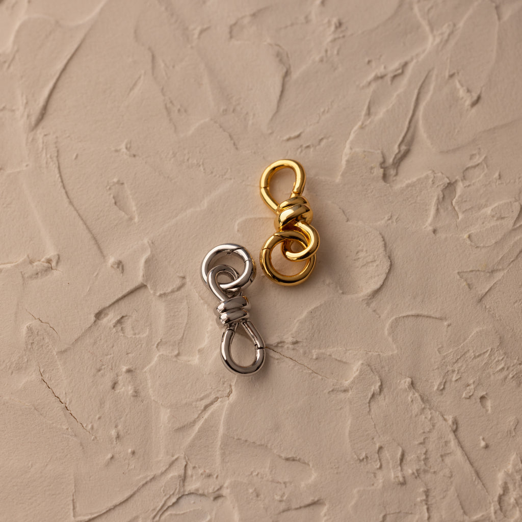 The Knot Charm Clasp, designed for secure fastening in gold and silver finishes, is displayed elegantly on a textured beige surface.