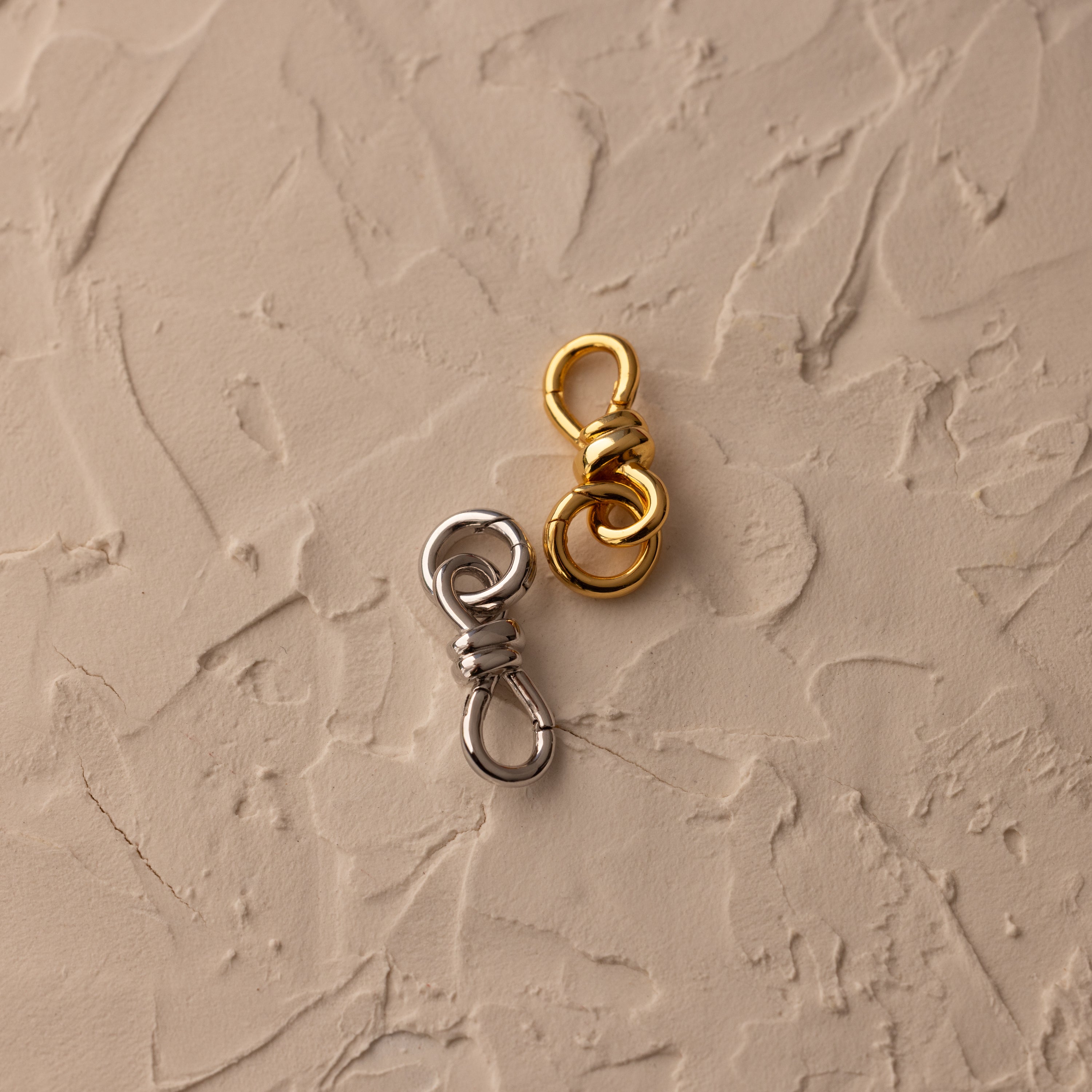 The Knot Charm Clasp, designed for secure fastening in gold and silver finishes, is displayed elegantly on a textured beige surface.