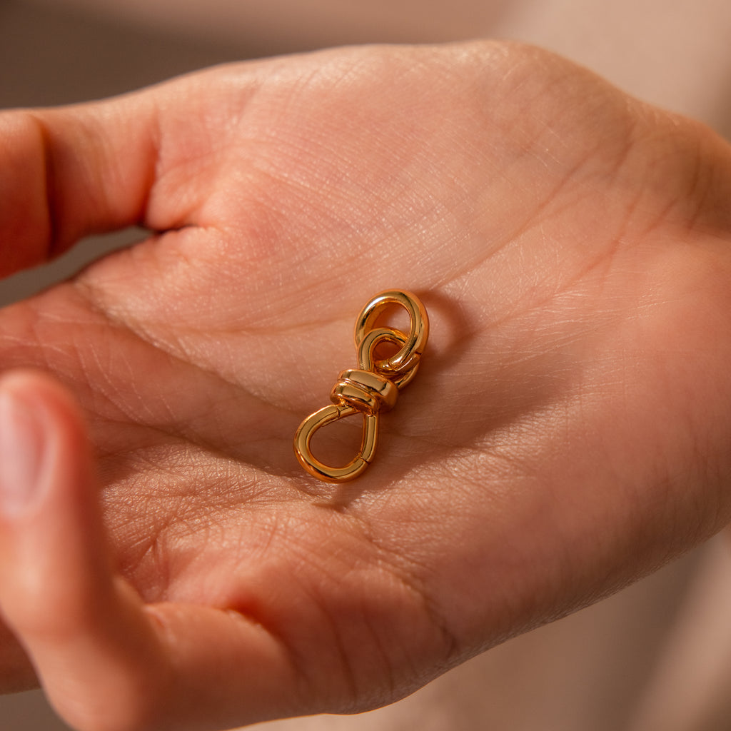 A hand holds the Knot Charm Clasp in 18K Gold, a small metal accessory featuring two loops—perfect for securely fastening your favorite jewelry pieces with style and elegance.