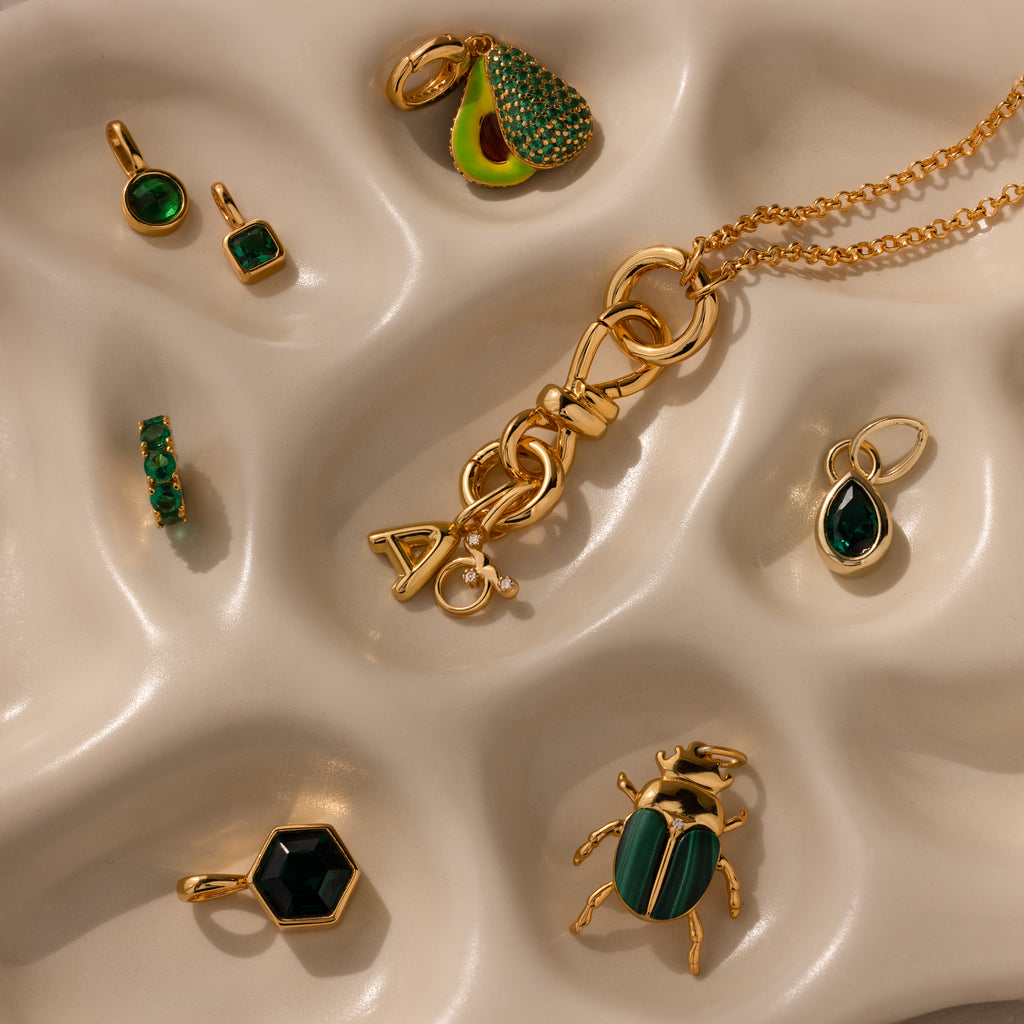 The Knot Charm Clasp in 18K Gold is showcased on a wavy beige surface, paired with gold and green jewelry pieces such as earrings, pendants, and a necklace.