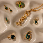 The Knot Charm Clasp in 18K Gold is showcased on a wavy beige surface, paired with gold and green jewelry pieces such as earrings, pendants, and a necklace.