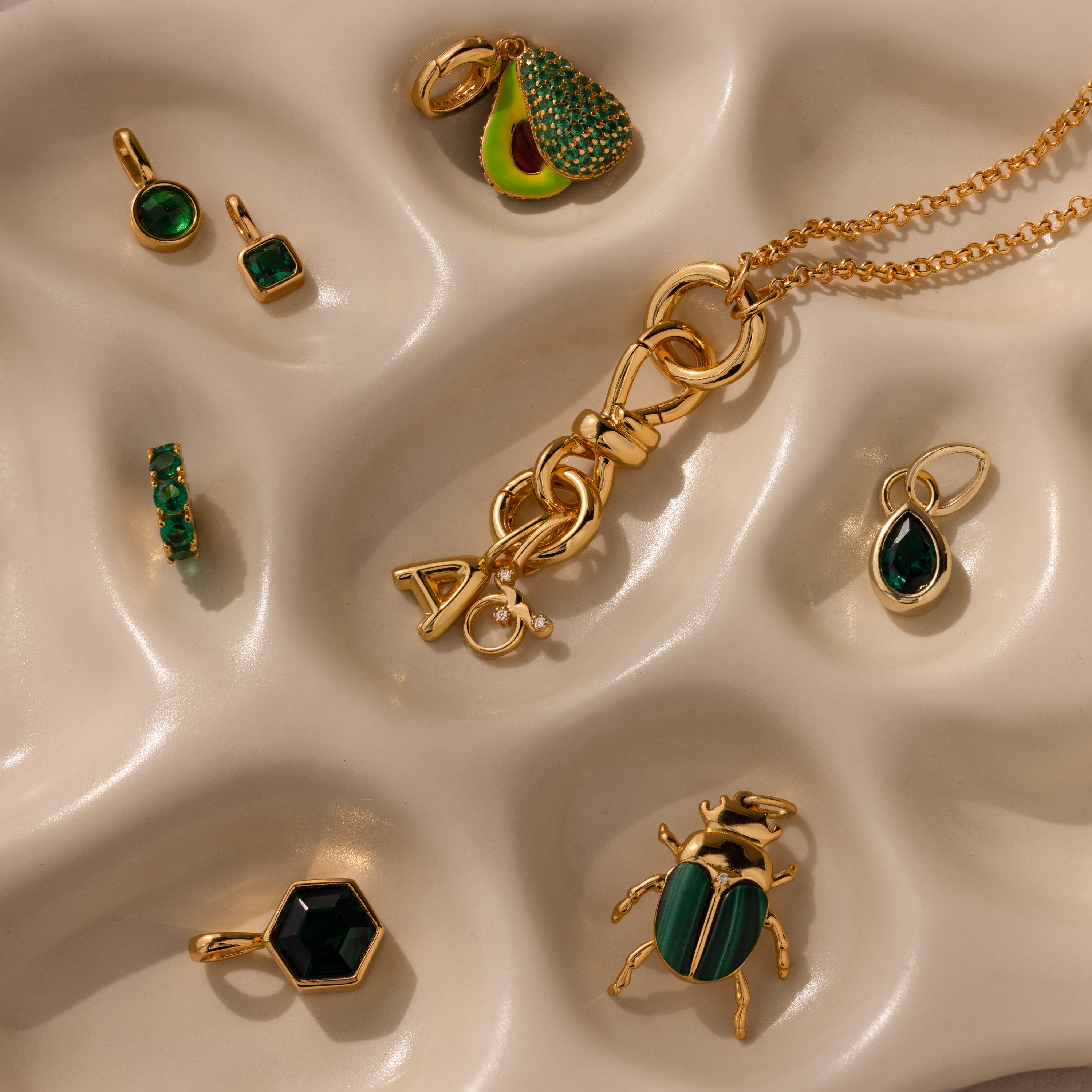 The Knot Charm Clasp in 18K Gold is showcased on a wavy beige surface, paired with gold and green jewelry pieces such as earrings, pendants, and a necklace.