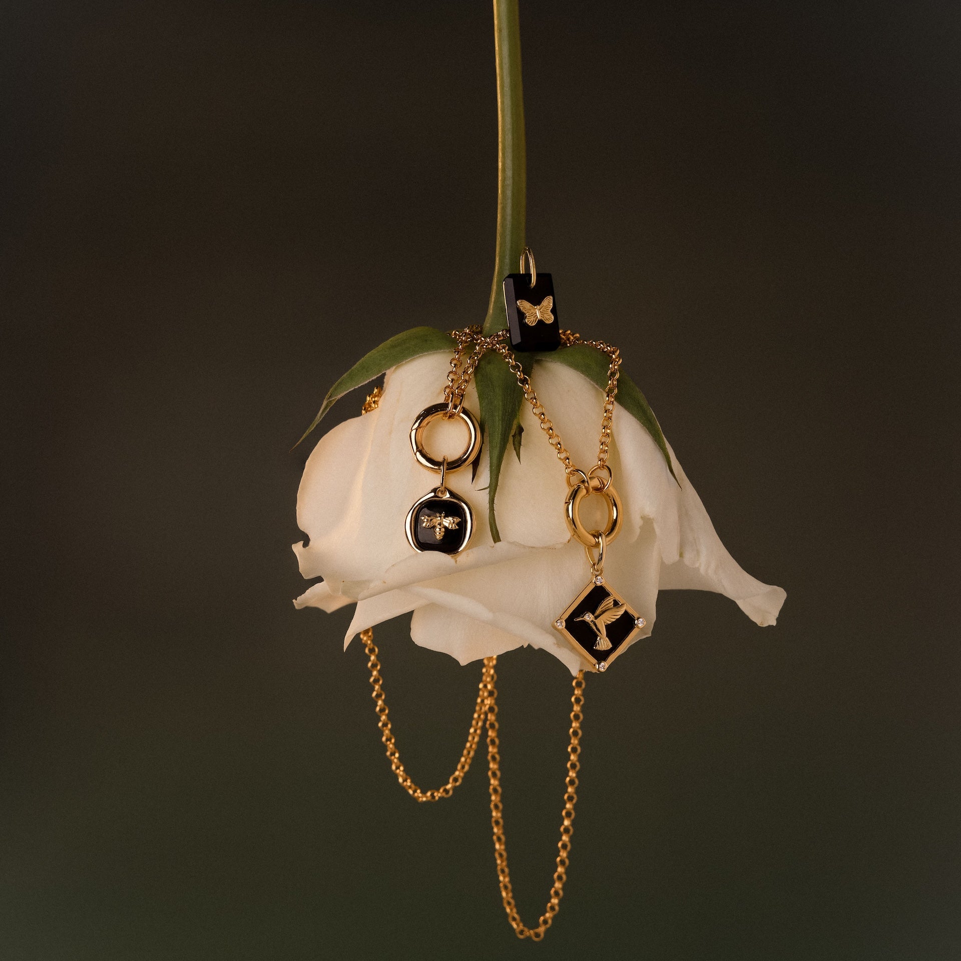 The Onyx Hummingbird Charm in 18K Gold is elegantly draped over an upside-down white rose on a dark background, creating a striking and delicate image that beautifully highlights this unique jewelry piece.
