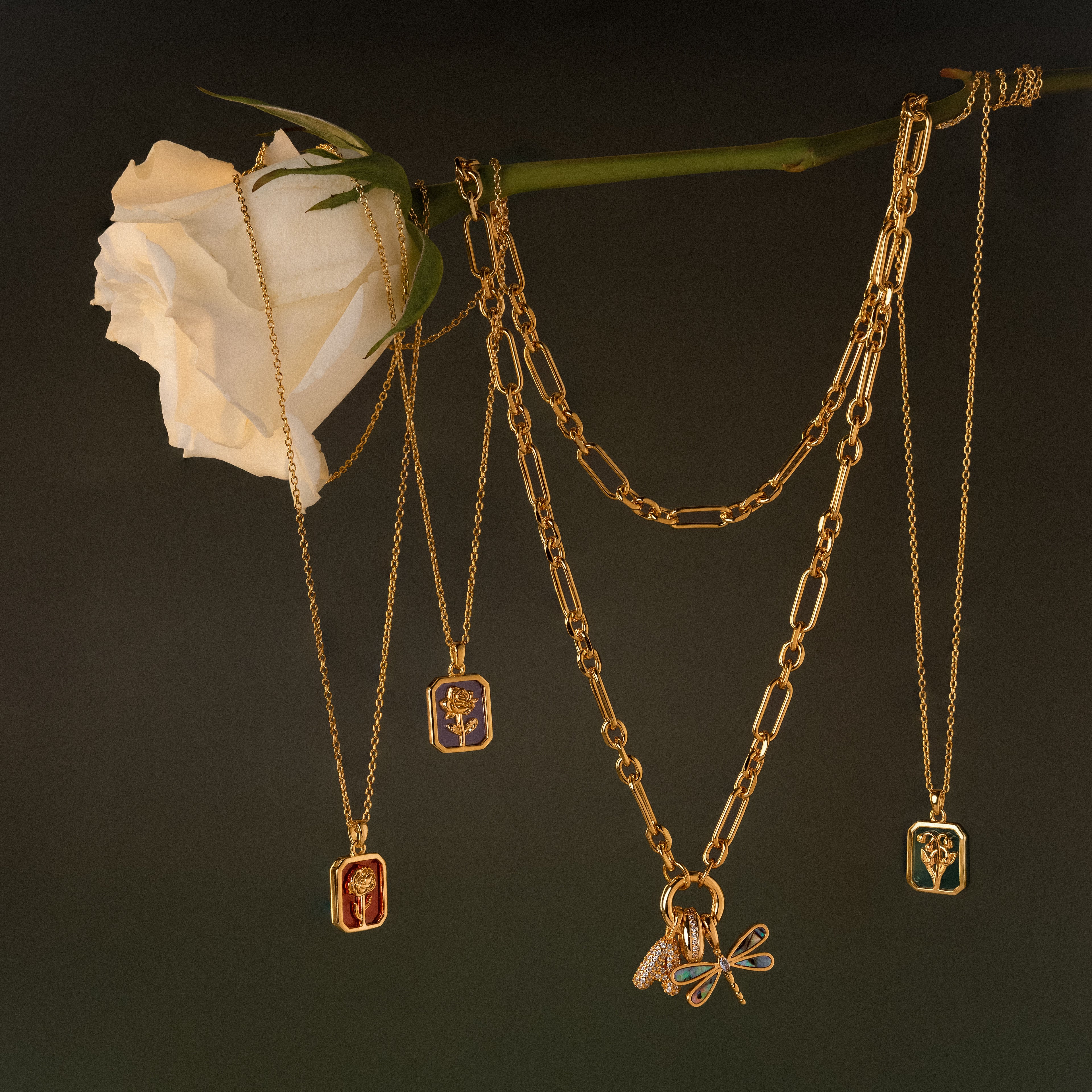 Birth Flower Charm Necklace pendants hang from a white rose stem against a dark background, creating an elegant product photo that highlights this unique jewelry piece.