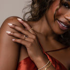 A woman in a satin dress displays manicured nails and gold jewelry, featuring the eye-catching Pave Garnet Ring, perfect for a January birthstone accessory.