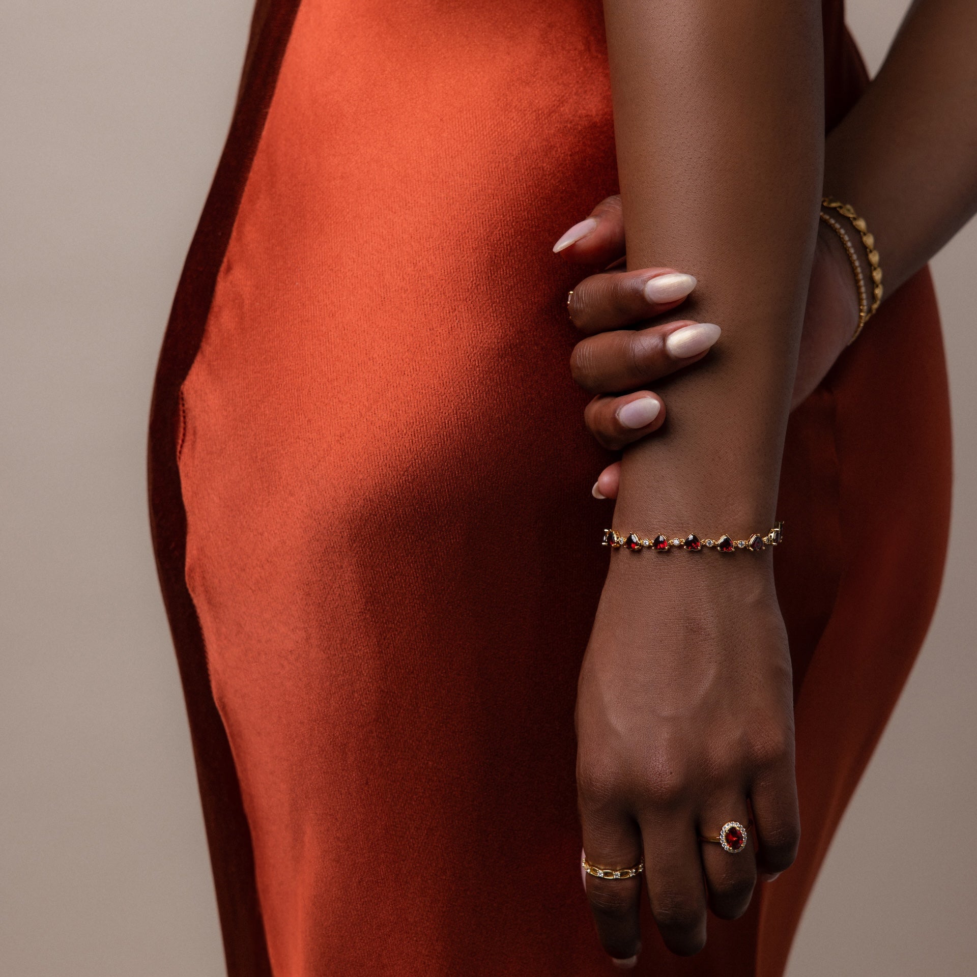 A woman in an orange satin dress showcases manicured nails and the Garnet Heart Tennis Bracelet in 18K Gold, featuring heart-shaped garnets, against a neutral background.