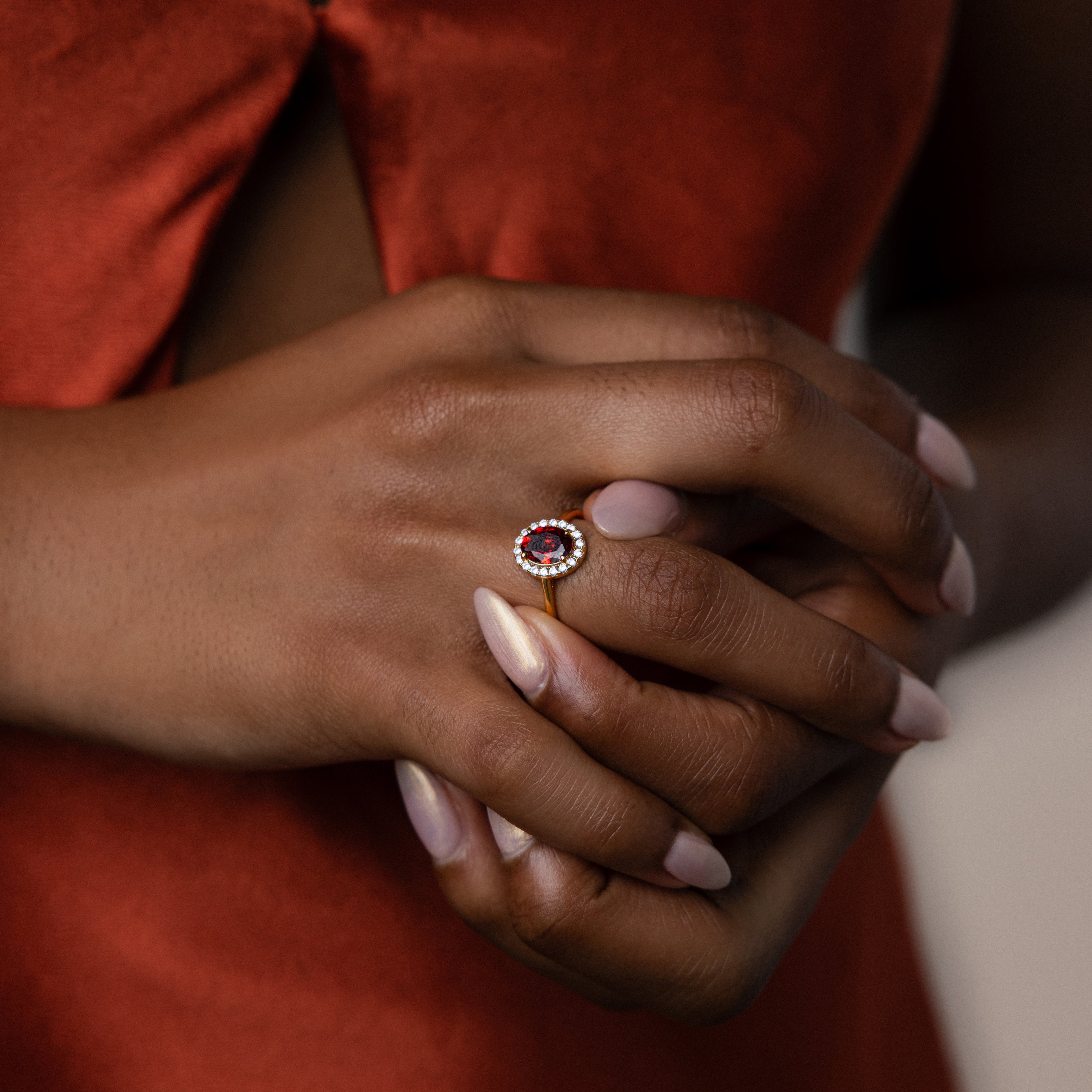 A person in a red dress displays the Pave Garnet Ring on their finger, hands gently clasped—a striking showcase of the January birthstone.