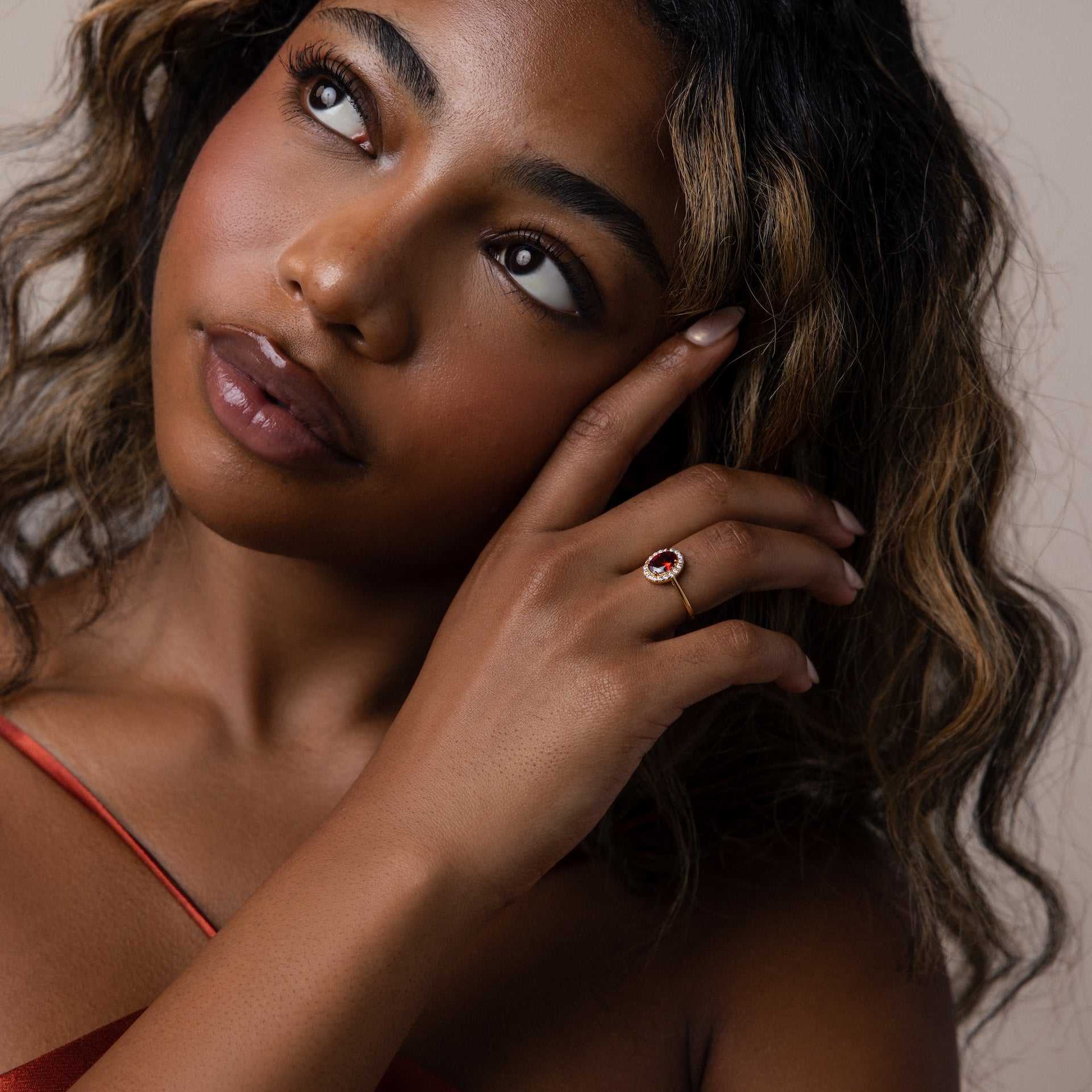 A woman with wavy hair poses in a red top, touching her face and showcasing the Pave Garnet Ring—her January birthstone—on her finger.