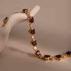 The Garnet Heart Tennis Bracelet in 18K Gold features red heart-shaped garnets and clear stones, shown draped over a white decorative hand display.