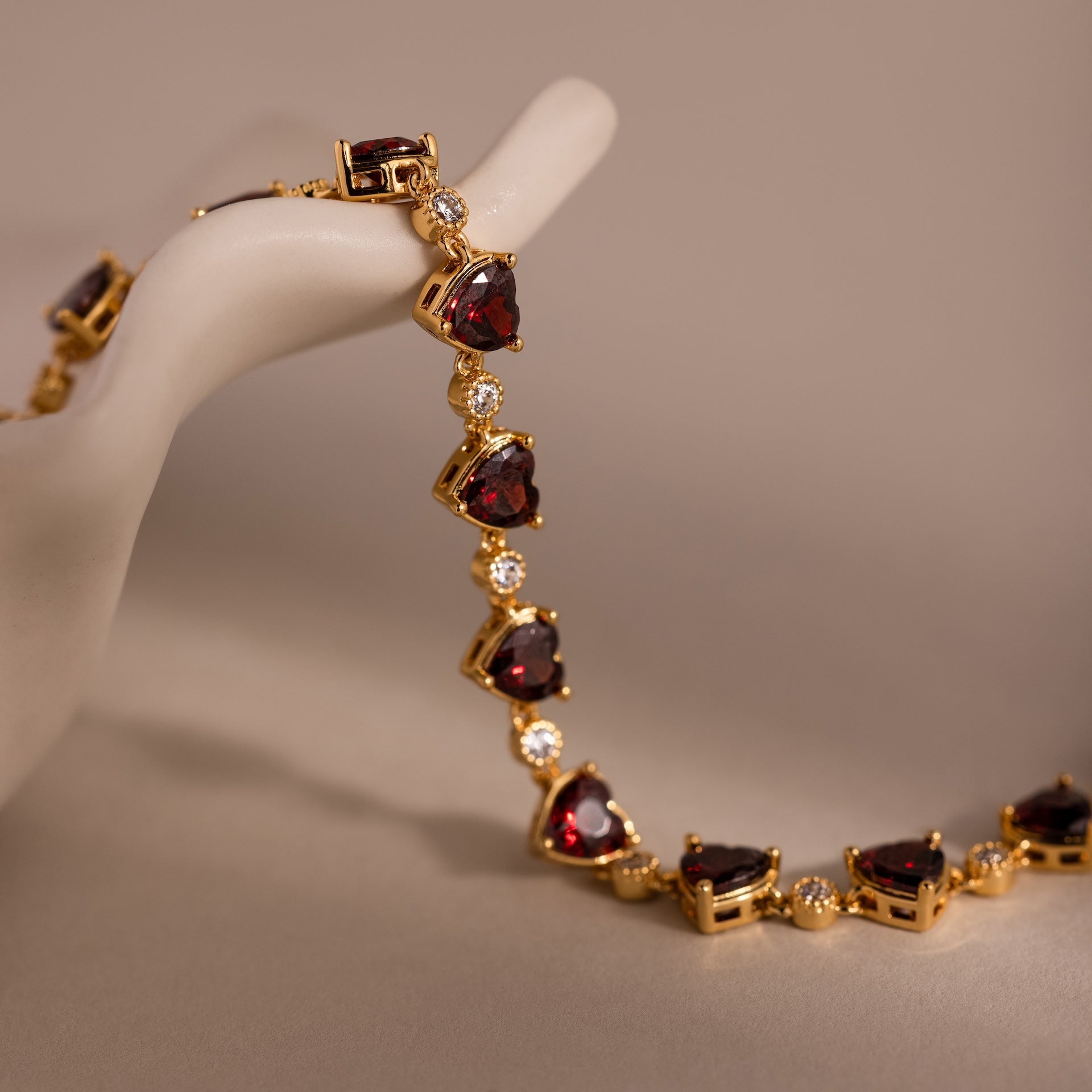 The Garnet Heart Tennis Bracelet in 18K Gold features red heart-shaped garnets and clear stones, shown draped over a white decorative hand display.