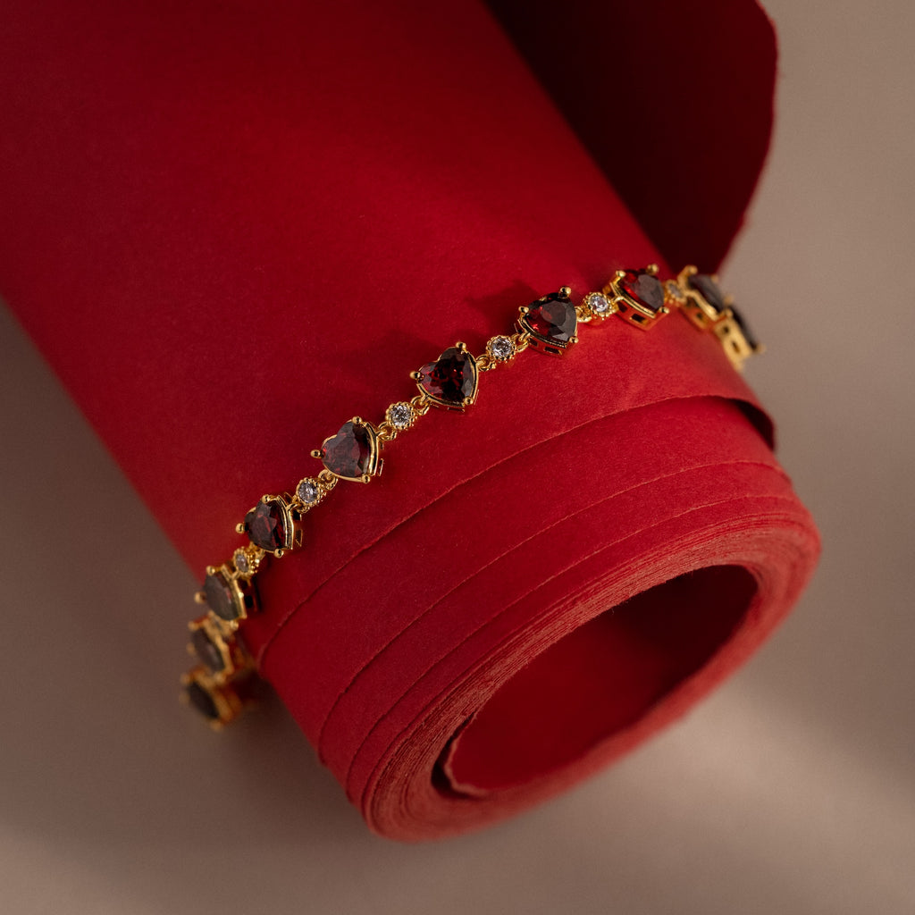 The Garnet Heart Tennis Bracelet in 18K Gold features heart-shaped garnets, elegantly displayed on rolled red paper with a beige background—a stunning statement piece for any occasion.