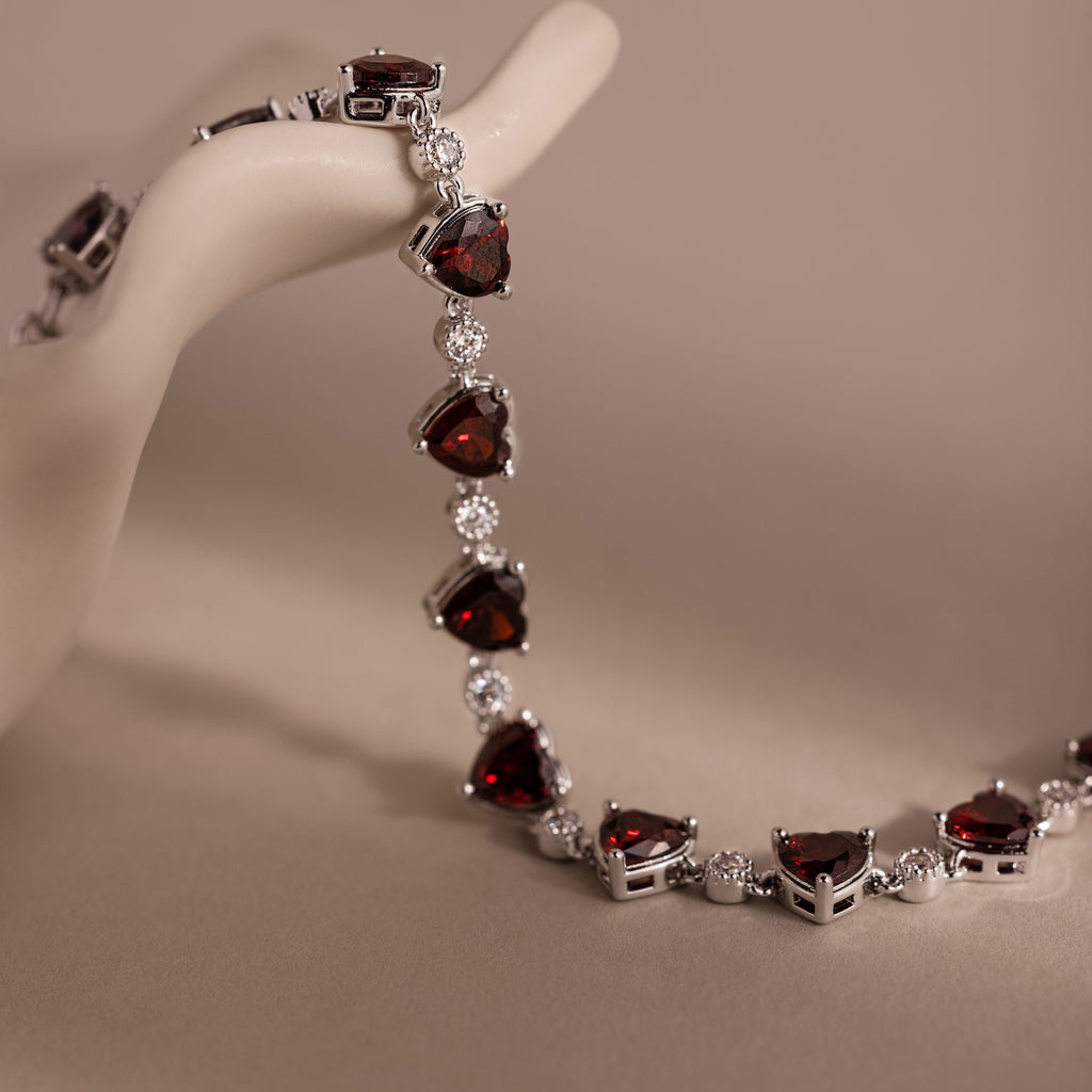 Garnet Heart Tennis Bracelet in Sterling Silver featuring red heart-shaped garnets and clear stones draped elegantly over a pale, sculpted hand display—a true statement piece.