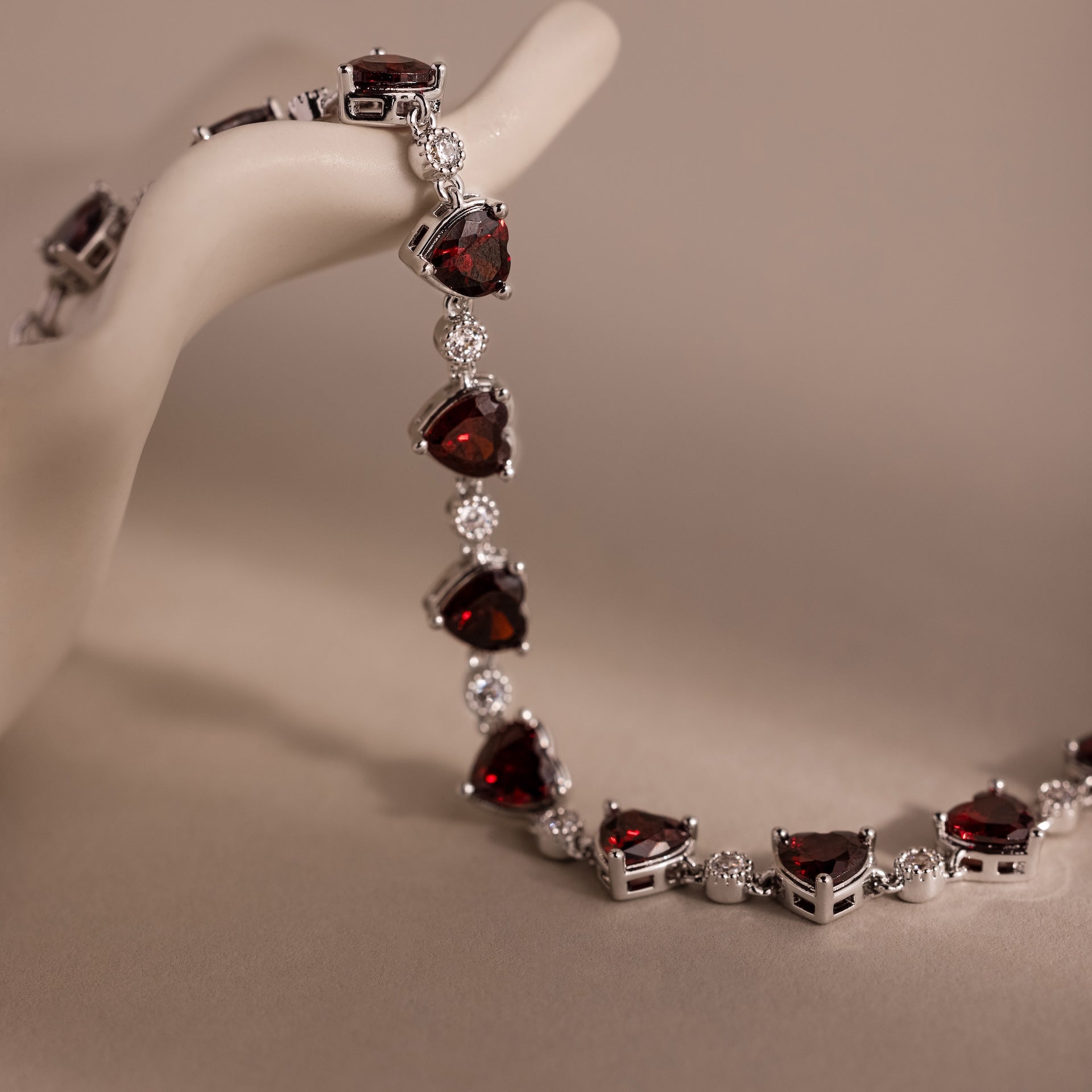 The Garnet Heart Tennis Bracelet features red heart-shaped garnets and clear stones, elegantly draped over a smooth, cream-colored display hand—a sophisticated statement piece.