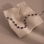 The Garnet Heart Tennis Bracelet in Sterling Silver features vivid red heart-shaped garnets on a draped, cream-colored textured surface—an elegant statement piece.