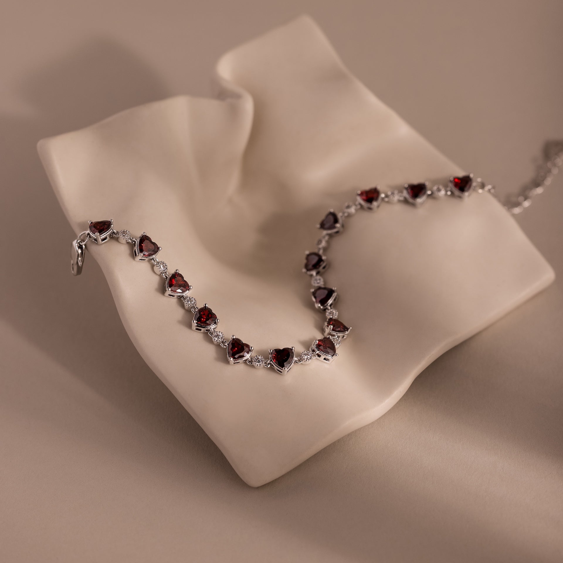 The Garnet Heart Tennis Bracelet in Sterling Silver features vivid red heart-shaped garnets on a draped, cream-colored textured surface—an elegant statement piece.