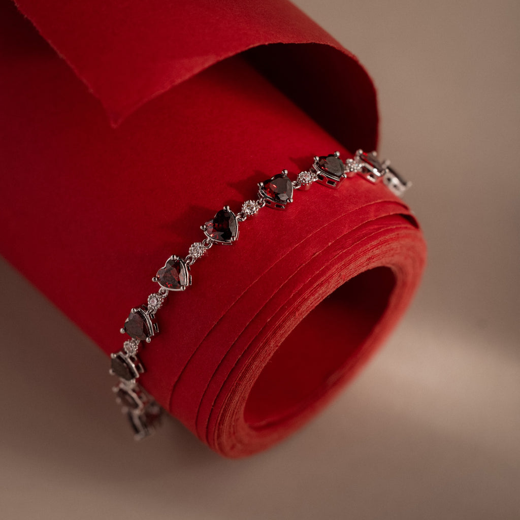 The Garnet Heart Tennis Bracelet in Sterling Silver features heart-shaped garnets and is displayed on a rolled red paper against a beige background, making it a striking statement accessory.