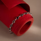 The Garnet Heart Tennis Bracelet in Sterling Silver features heart-shaped garnets and is displayed on a rolled red paper against a beige background, making it a striking statement accessory.