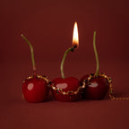 Three cherries, one with a burning stem, are draped with the Garnet Heart Tennis Bracelet in 18K Gold—its heart-shaped garnets glowing against a vibrant red background.