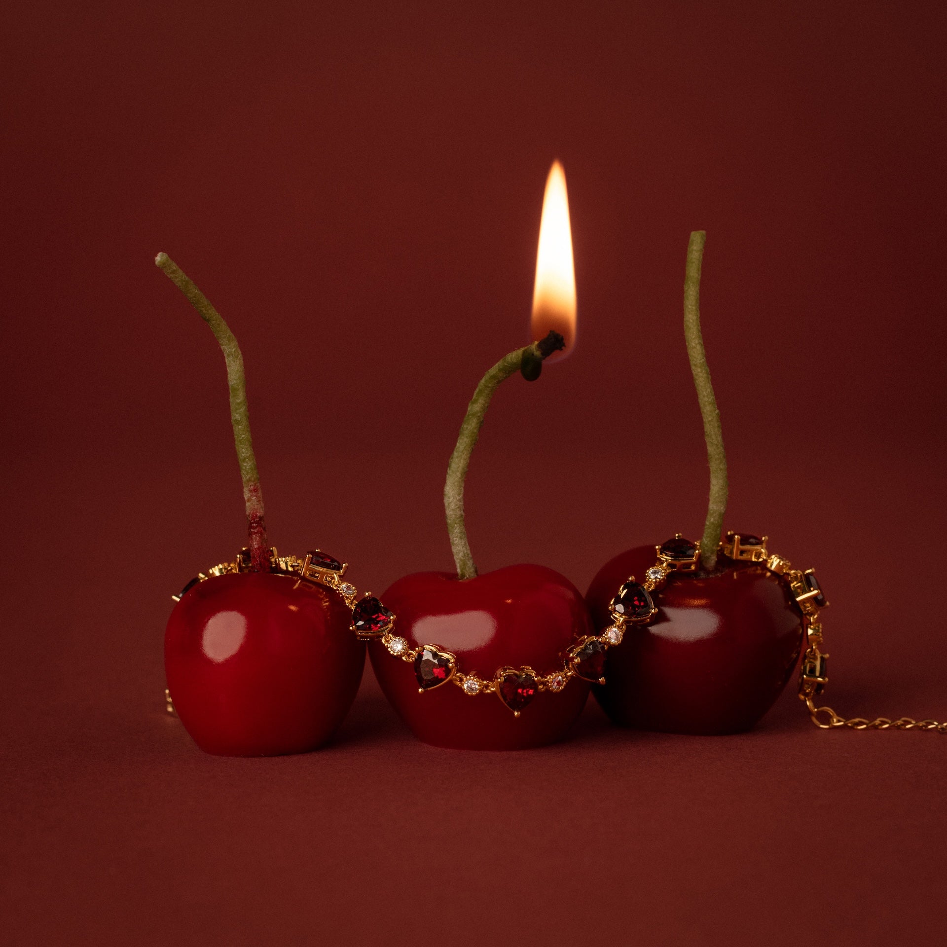 Three cherries, one with a burning stem, are draped with the Garnet Heart Tennis Bracelet in 18K Gold—its heart-shaped garnets glowing against a vibrant red background.