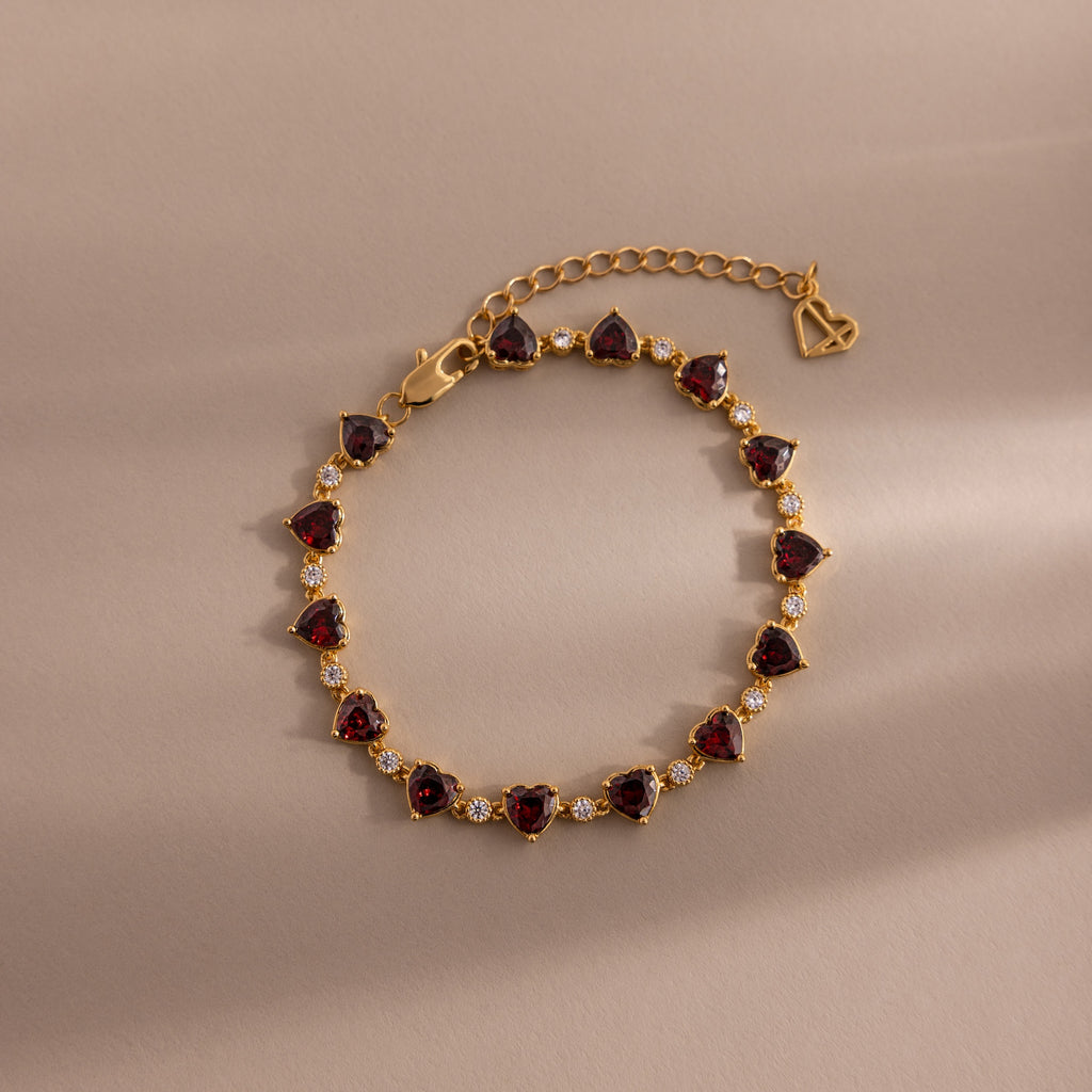 The Garnet Heart Tennis Bracelet in 18K Gold features red heart-shaped garnets, clear stones, an adjustable chain, and a heart charm—perfect as a standout statement bracelet.