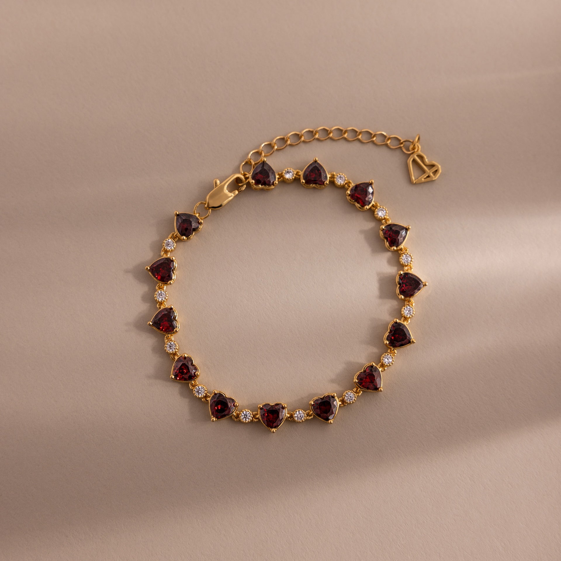The Garnet Heart Tennis Bracelet in 18K Gold features red heart-shaped garnets, clear stones, an adjustable chain, and a heart charm—perfect as a standout statement bracelet.