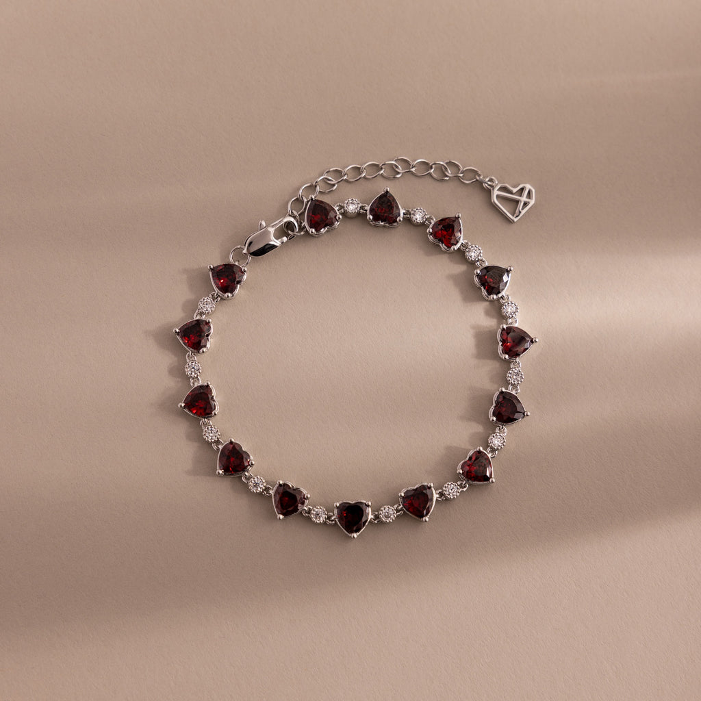 The Garnet Heart Tennis Bracelet in Sterling Silver features heart-shaped garnets and sparkling white stones, making it a standout statement piece against a beige background.