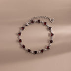The Garnet Heart Tennis Bracelet in Sterling Silver features heart-shaped garnets and sparkling white stones, making it a standout statement piece against a beige background.