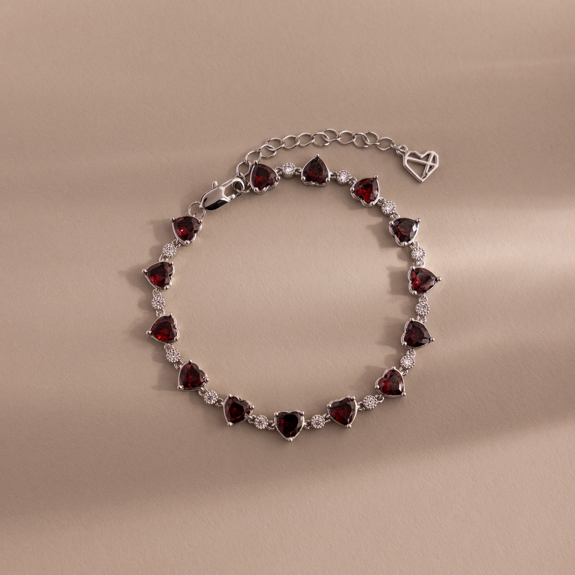 The Garnet Heart Tennis Bracelet in Sterling Silver features heart-shaped garnets and sparkling white stones, making it a standout statement piece against a beige background.