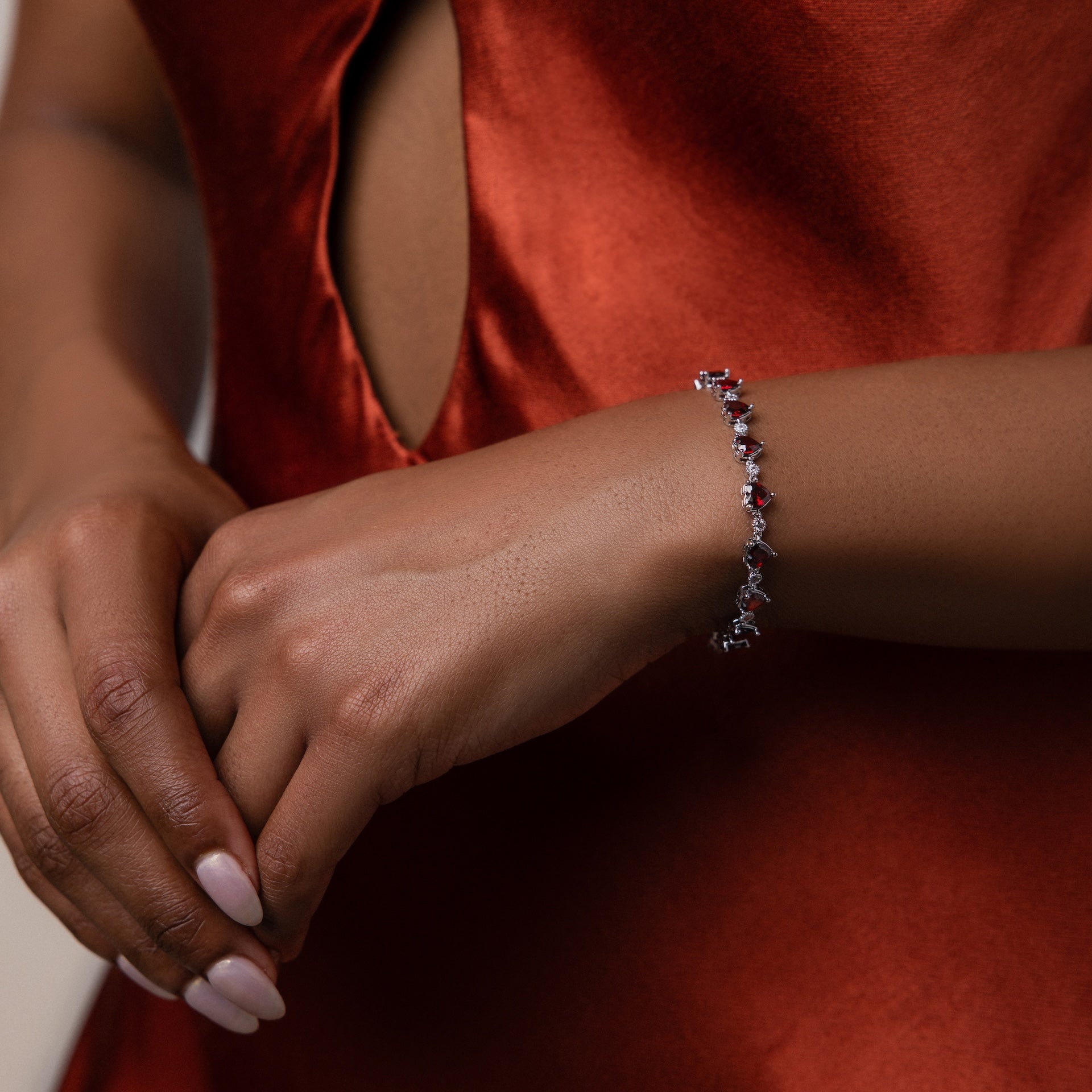 Woman in a red satin dress wears the Garnet Heart Tennis Bracelet in Sterling Silver, featuring delicate heart-shaped garnets and white gemstones—a striking statement piece.