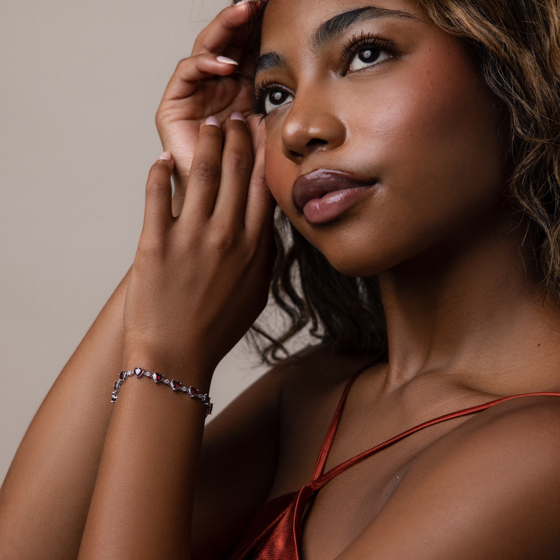 A woman in a red dress thoughtfully touches her face, showcasing the Garnet Heart Tennis Bracelet with heart-shaped garnets against a neutral background.