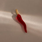 The Chili Pepper Charm in 18K Gold features a red chili pepper-shaped pendant with a gold loop, shown on a softly lit, neutral surface—a playful addition to any jewelry collection.