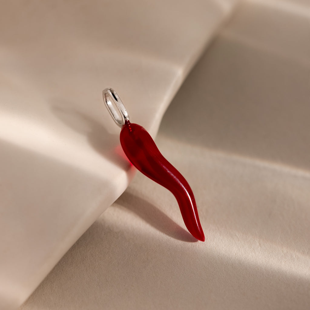 The Chili Pepper Charm in Sterling Silver features a vibrant red design with a silver loop, resting on a soft beige surface—a bold and stylish addition to your jewelry collection.
