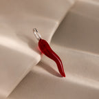 The Chili Pepper Charm in Sterling Silver features a vibrant red design with a silver loop, resting on a soft beige surface—a bold and stylish addition to your jewelry collection.