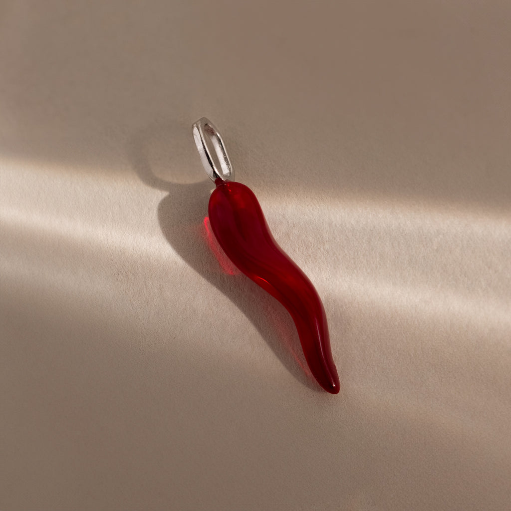 The Chili Pepper Charm in Sterling Silver features a red glass chili pendant with a silver loop, ideal for charm necklaces, shown on a neutral background with gentle lighting and shadows.