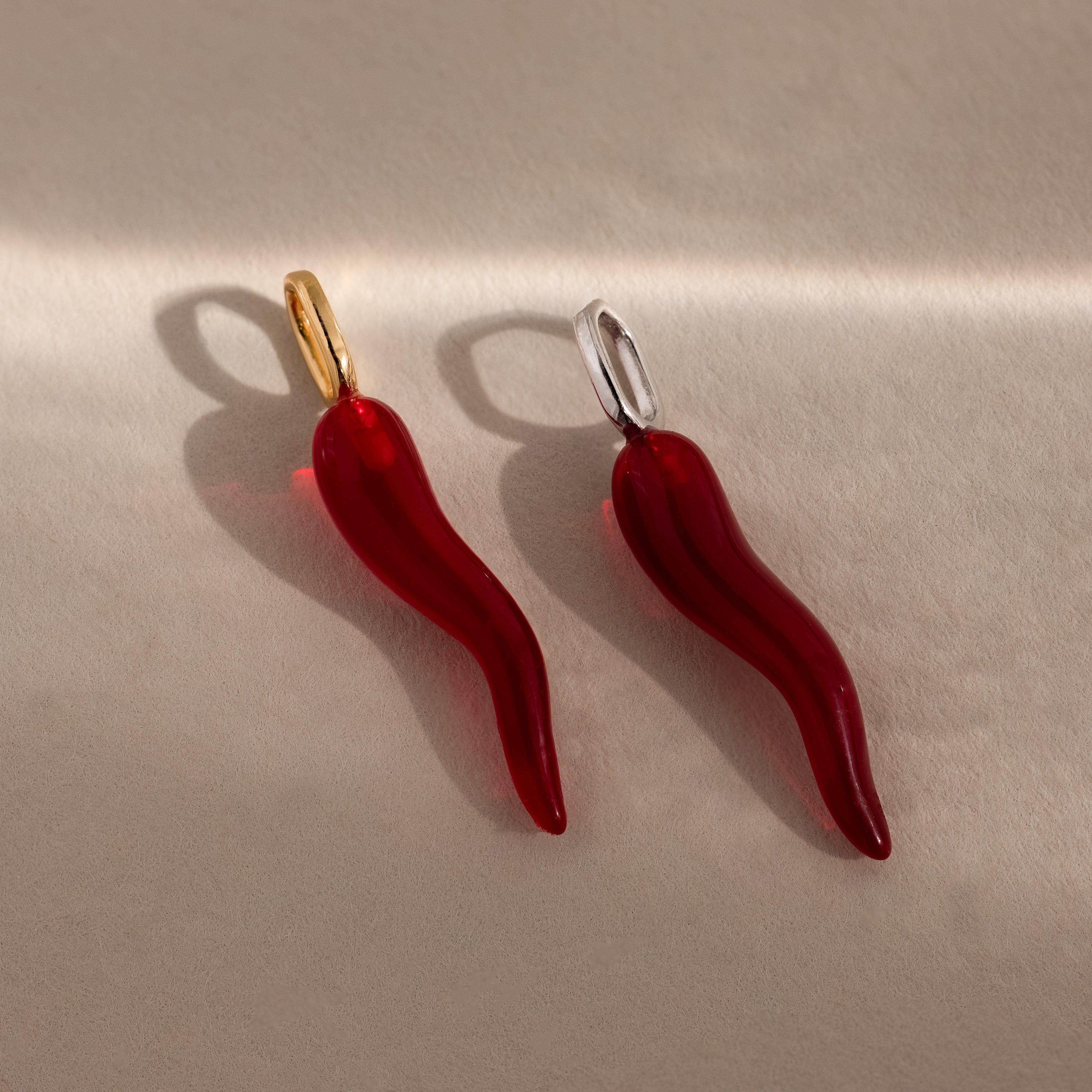 Two Chili Pepper Charm pendants with gold and silver loops rest on a beige surface, casting delicate shadows—ideal for adding a unique accent to your jewelry or as a charm necklace addition.