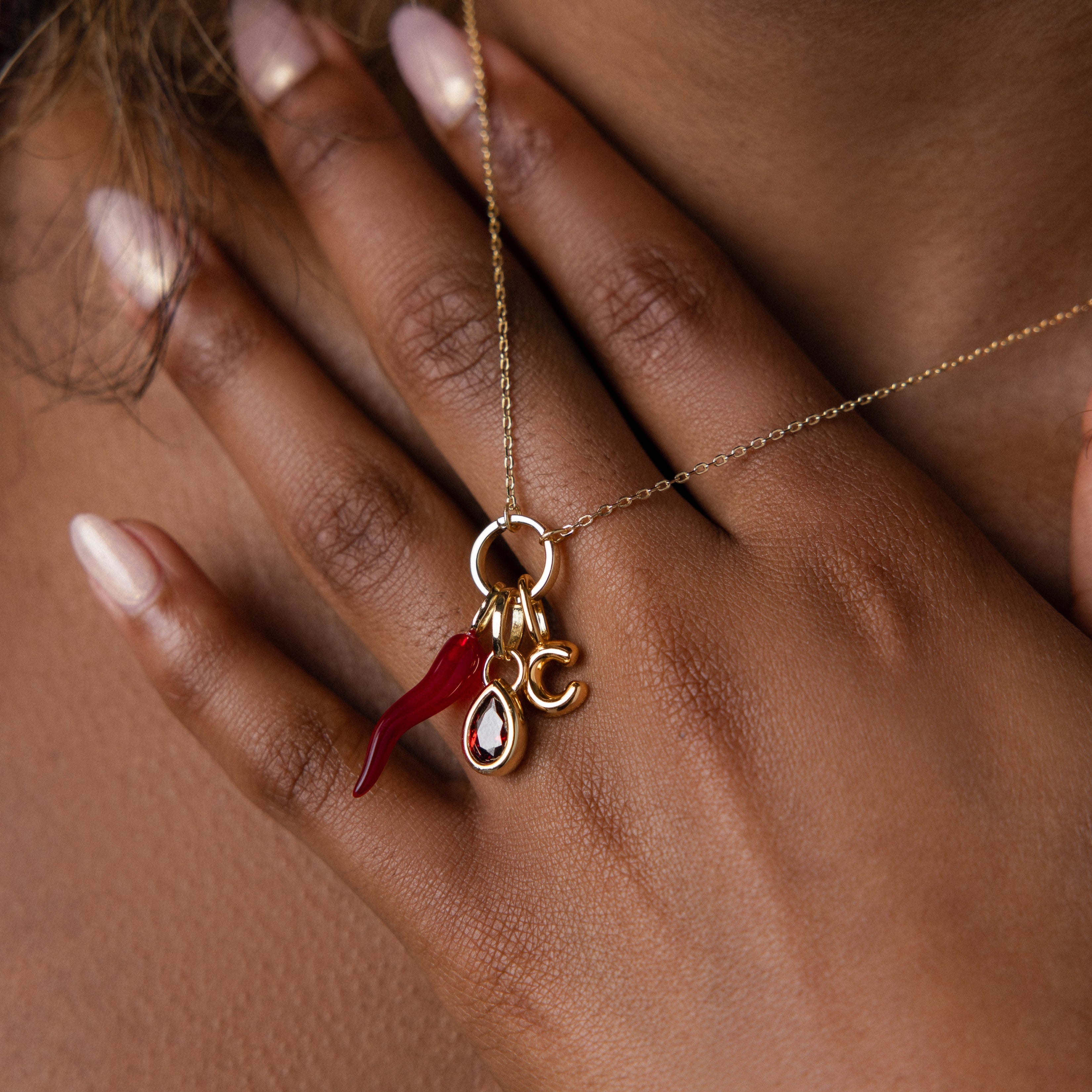 A hand with manicured nails holds a necklace featuring the Chili Pepper Charm, a sparkling gem, and the letter C—a distinctive piece of jewelry bursting with personality.