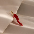 The Chili Pepper Charm in 18K Gold, featuring a red design with a gold loop, rests on a soft beige surface—a striking addition to any charm necklace or jewelry collection.