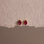 Rhombus Ruby Flatback Studs feature small, diamond-shaped red gemstones for understated chic. These modern earrings add a touch of glamour to any look, shown here against a textured beige background.