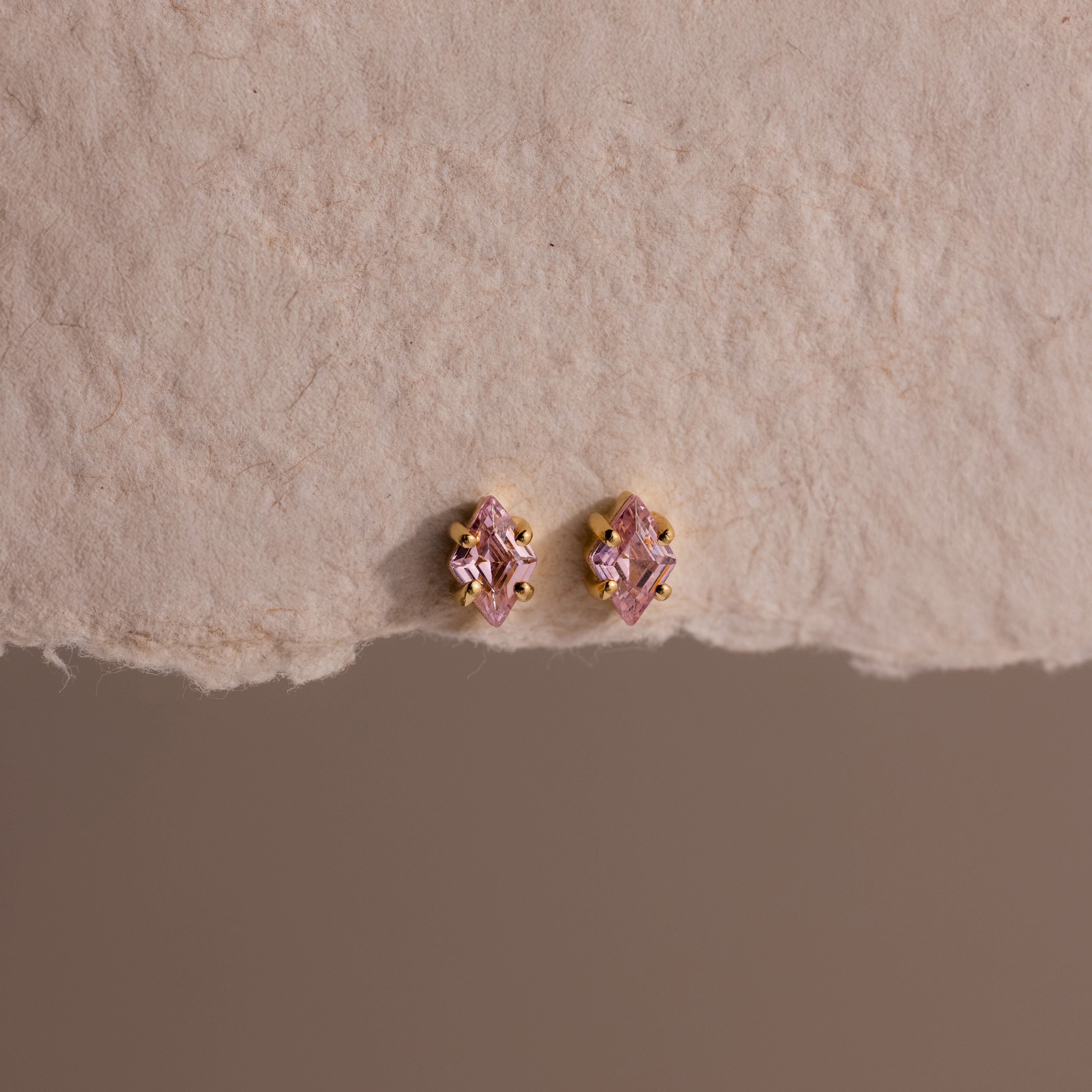 The Rhombus Pink Tourmaline Flatback Studs add modern glamour with their small pink stones and gold settings on a textured beige surface.
