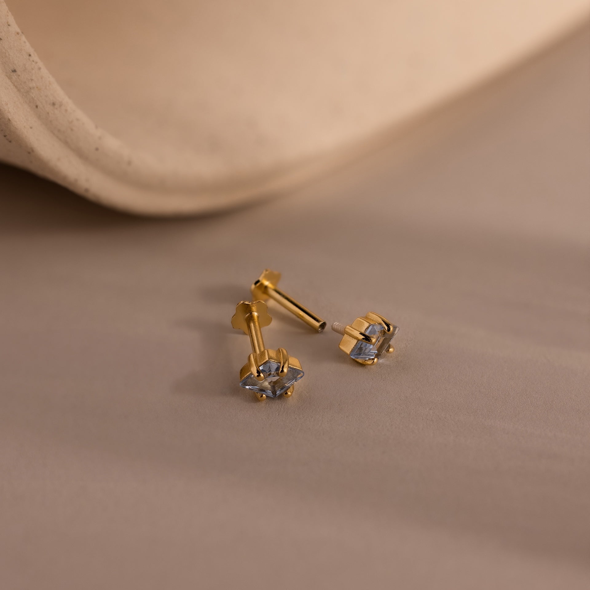 Rhombus Blue Zircon Flatback Studs feature square-cut blue zircon stones for a modern, understated chic look, shown on a soft beige surface with a gently curved background.