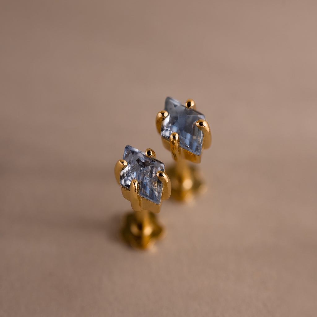 The Rhombus Birthstone Flatback Studs feature rectangular blue gemstones set in gold, adding modern glamour to your look. Shown on a beige surface, these studs bring understated chic to any jewelry collection.