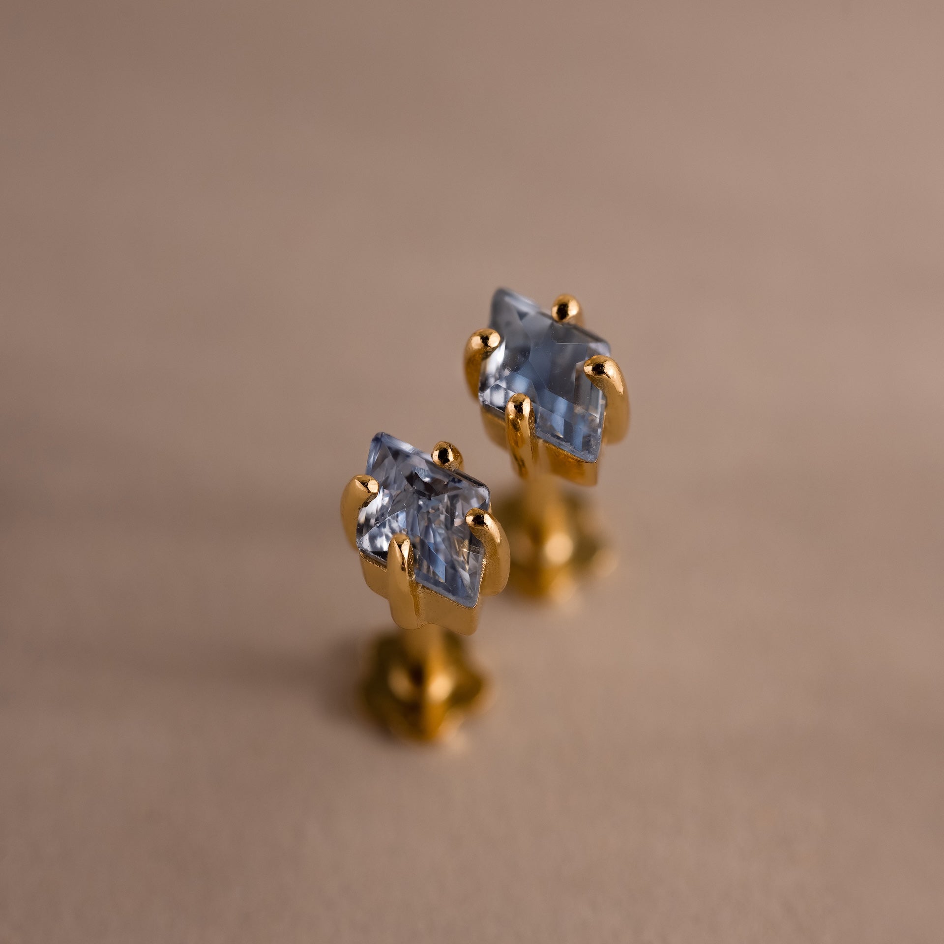 The Rhombus Birthstone Flatback Studs feature rectangular blue gemstones set in gold, adding modern glamour to your look. Shown on a beige surface, these studs bring understated chic to any jewelry collection.
