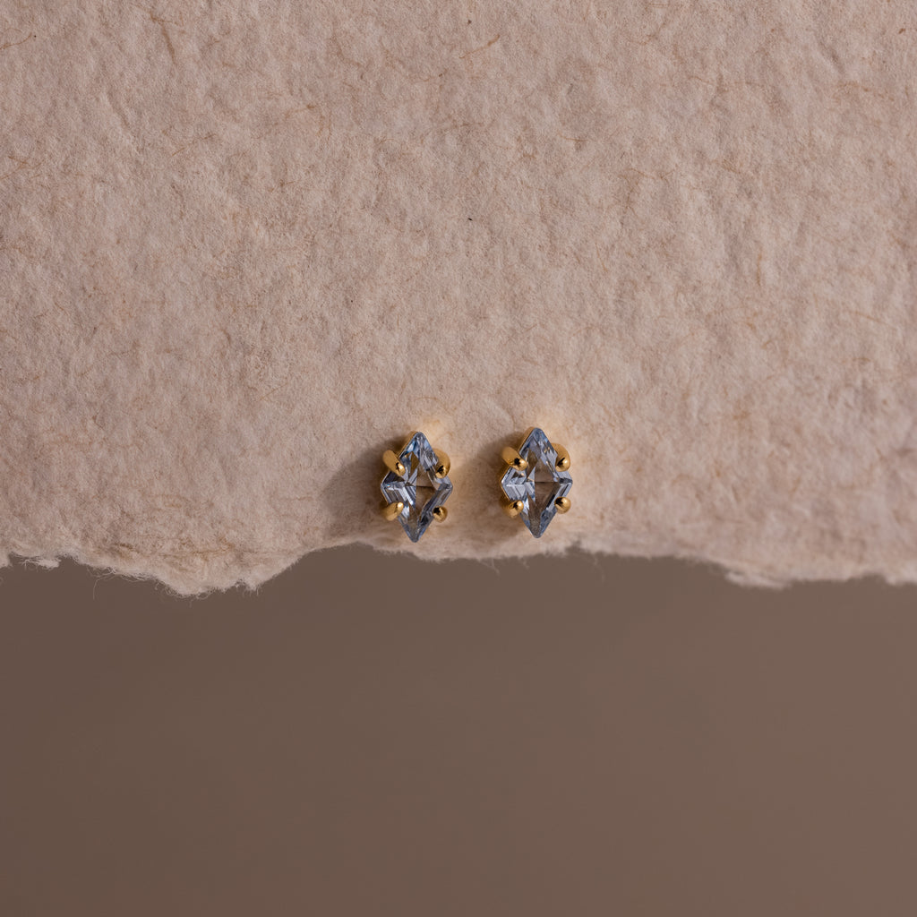 A pair of small, gold and silver stud earrings displayed on textured beige paper, these Rhombus Aquamarine Flatback Studs exude understated chic and modern glamour.