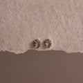 A pair of small, gold and silver stud earrings displayed on textured beige paper, these Rhombus Aquamarine Flatback Studs exude understated chic and modern glamour.
