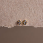 A pair of small, gold and silver stud earrings displayed on textured beige paper, these Rhombus Aquamarine Flatback Studs exude understated chic and modern glamour.