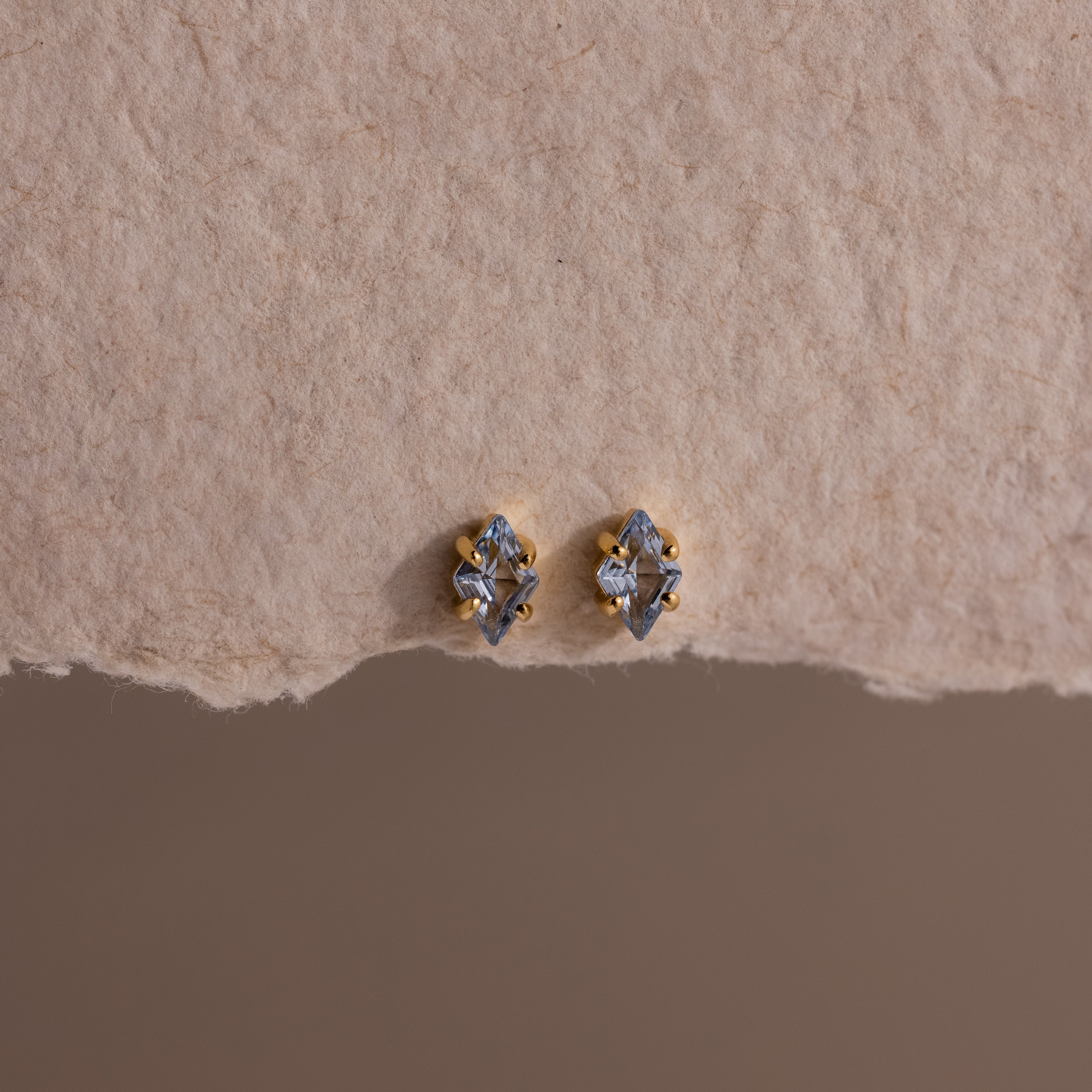 A pair of small, gold and silver stud earrings displayed on textured beige paper, these Rhombus Aquamarine Flatback Studs exude understated chic and modern glamour.
