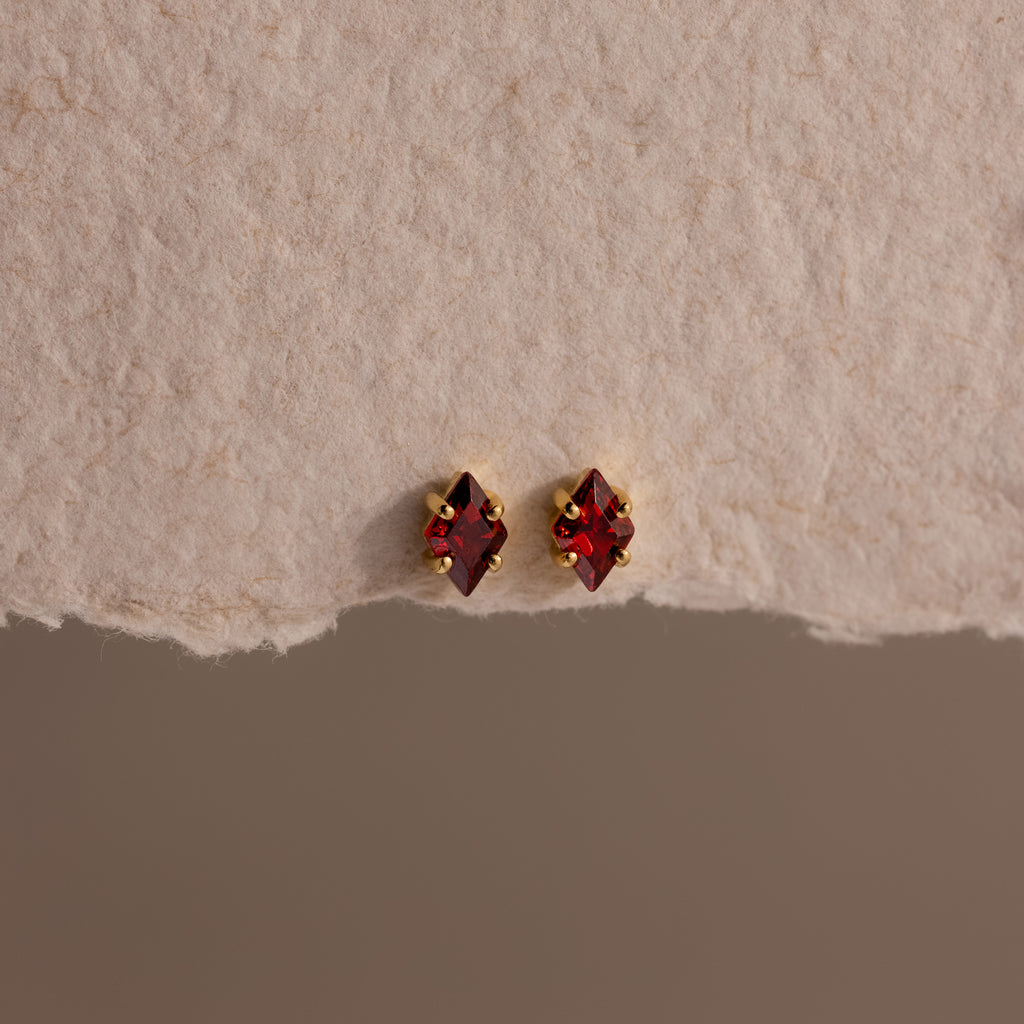 The Rhombus Garnet Flatback Studs, featuring small red gemstones in gold settings, add subtle jewelry charm on a textured off-white surface.