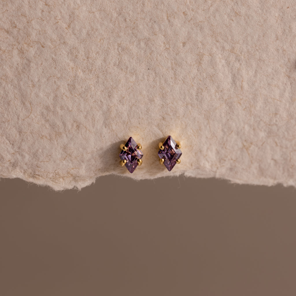 The Rhombus Amethyst Flatback Studs feature small diamond-shaped purple stones with gold accents, adding modern glamour to a chic look. Displayed on a textured beige surface.