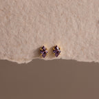 The Rhombus Amethyst Flatback Studs feature small diamond-shaped purple stones with gold accents, adding modern glamour to a chic look. Displayed on a textured beige surface.